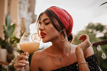 Attractive brunette haired girl with pink headband, round silver earrings, bright bandana in black shirt drinking cocktail..