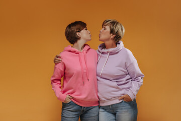 Fashionable two women with short modern hairstyle in trendy bright pink hoodies and cool jeans hugging and blowing kisses on orange backdrop..