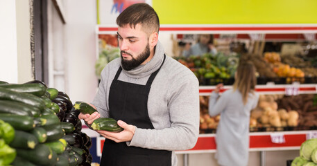 Male shop assistant lays fresh cucumber on counter in grocery shop. High quality photo