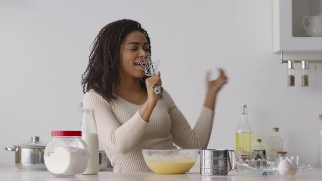 Young positive african american woman singing into whisk and dancing at kitchen while cooking dough for bakery