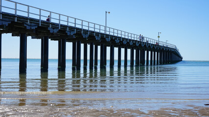 Fototapeta premium Looking up at long wooden pier, with blue sky, calm water, gentle waves