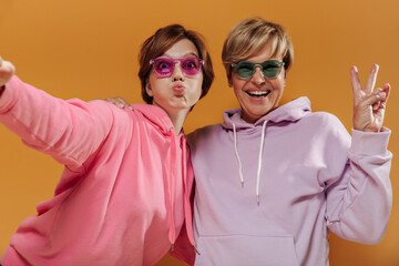 Funny two women with short modern hairstyle in bright cool sunglasses and pink and lilac hoodie taking selfie and having fun on orange backdrop.. - Powered by Adobe