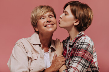 Young short haired girl in plaid red shirt kissing on cheek her grandmother with blonde hair in beige jacket on pink backdrop..