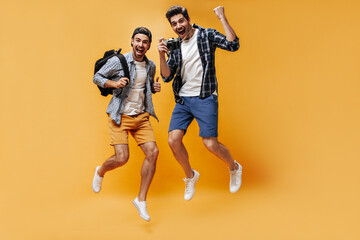 Fototapeta na wymiar Cool happy men in shorts and checkered shirts jump and rejoice on orange background. Happy guys hold retro camera and backpack.