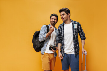 Handsome men talk on orange background. Cool young travelers in checkered shirts pose with backpack, suitcase and retro camera on orange background.