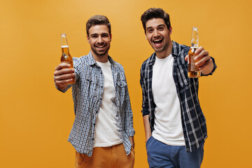 Excited young men in white trendy t-shirts and checkered shirts rejoice and hold beer bottles on...