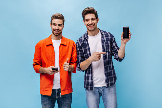 Cool young excited men in stylish outfits pose on blue background. Guy in orange jacket shows thumb up. Man in checkered shirt points at phone screen.