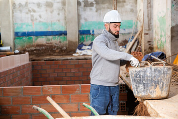 Young bearded bricklayer installing brick wall in building under construction..