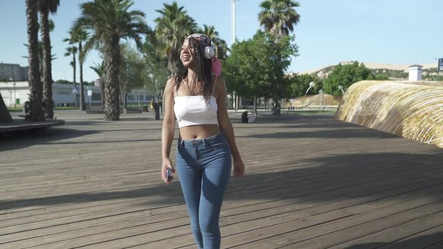 Joyful hispanic girl with afro hairs dancing outdoors in city in front of palm trees during beautiful weather in nature. Listen music on headphones.