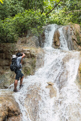 Naklejka na ściany i meble Young man with backpack in waterfall in mountain taking photos and hikign 