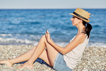  Young woman in a hat with a phone on the beach near the sea is making a selfie. Resting, traveling and relaxing concept. Copy space
