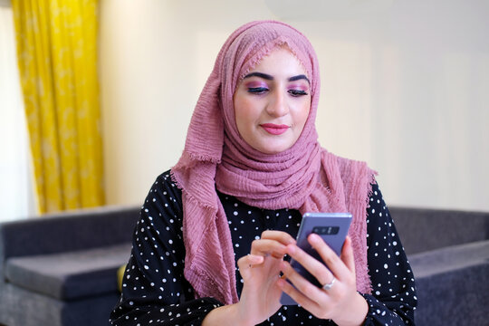 Online Buying Concept Of A Arabic Woman Wearing Head Scarf As Part Of Traditional Custome Using Mobile Cell Phone.  Emirati Lady Using Mobile Device Browsing The Internet Or Doing Online Banking