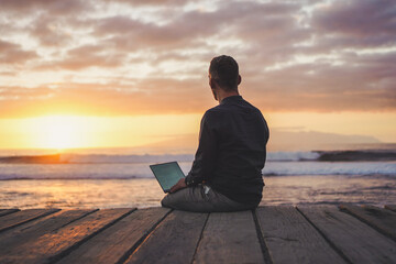 back of a sitting digital nomad remote working man on a boardwalk at the beach working with laptop during sunset

