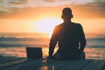 back of a sitting digital nomad remote working man on a boardwalk at the beach working with laptop during sunset
