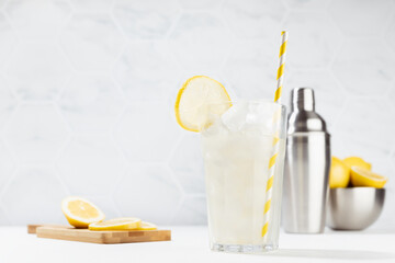 Fresh summer classic lemonade in glass with straw, ice, ingredients - lemons in silver bowl, shaker, slices fruit on board in soft light modern white kitchen interior with marble tile in sunny day.