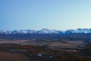 Awesome landscape with mountain river serpentine in valley among hills and forest in autumn colors with view to great snowy mountain range in sunset. High snow-covered mountains and autumn valley.