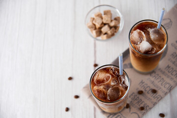 Iced coffee in a tall glass with cream or milk and beans, straws on light background. Cold tasty summer refreshment beverage concept. Selective focus, copyspace. Flat lay, top view.