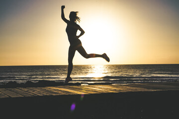 Fototapeta na wymiar silhouette of a successful, strong and sportive female doing champion pose with fists on beach boardwalk during sunset or sunrise 