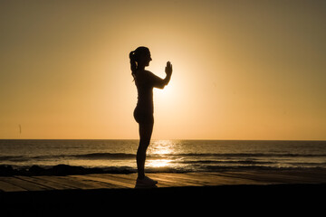 Fototapeta premium silhouette of a sportive female in namaste pose practicing yoga & meditation on beach boardwalk during sunset or sunrise 