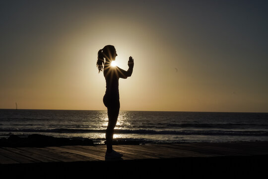 silhouette of a sportive female in namaste pose practicing yoga & meditation on beach boardwalk during sunset or sunrise 