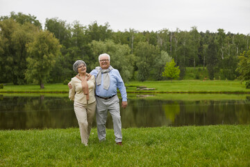 Naklejka na ściany i meble An elderly couple in casual clothes are walking outdoors, hugging each other and against the background of the forest and the river.