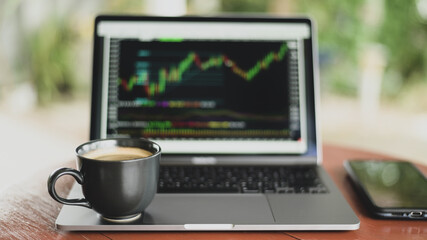 Fototapeta premium Coffee mug placed on laptop with stock graph on screen.