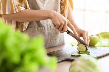 Young woman cutting cucumber in kitchen, closeup