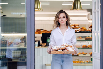 Tight lipped woman posing with a tray full of pastry in a doorstep of her shop
