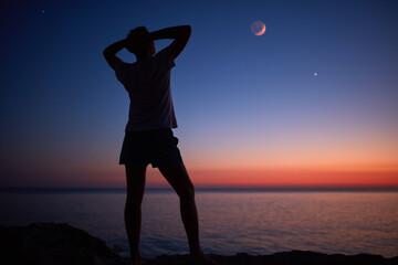 Silhouette of a man looking at the Moon and stars over sea ocean horizon.