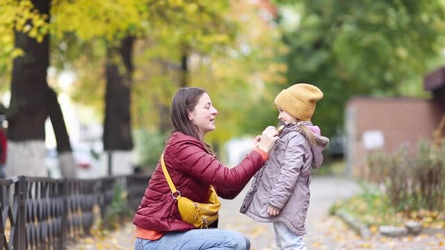 family mother with child on street of city autumn. mom helps her daughter to button up her jacket in fall.