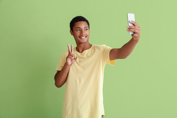 Happy African-American teenage boy taking selfie on color background