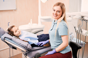 Obraz premium Adorable kid looking at camera and smiling while lying in dental chair. Woman dentist in sterile gloves sitting beside cute little girl in dental office. Concept of pediatric dentistry and dental care