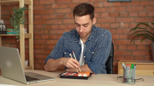 Handsome male freelancer eating sushi rolls while reading news on laptop in desk at home office. Happy Caucasian young bearded man watching laptop computer and eating Japanese food in living room.