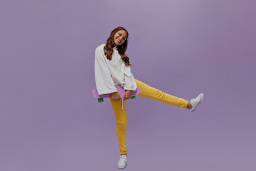 Full-length portrait of active teenager holding longboard. Pretty young girl in white hoodie and yellow pants poses with skateboard on purple background.