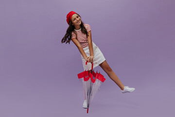 Attractive young girl poses with umbrella on purple background. Joyful teenager in red beret, white skirt and striped t-shirt smiles on isolated.