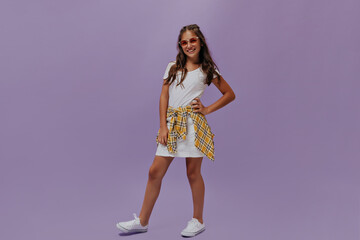 Curly tanned teenager poses on purple background. Attractive young girl in white t-shirt and skirt smiles sincerely on isolated.