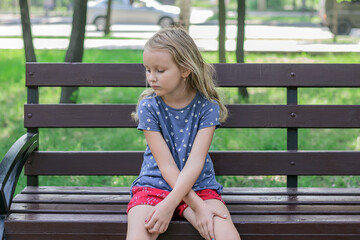 little girl, blonde, sitting sad in the park on a bench, crossing her arms, portrait of a child with a pensive look, outdoors