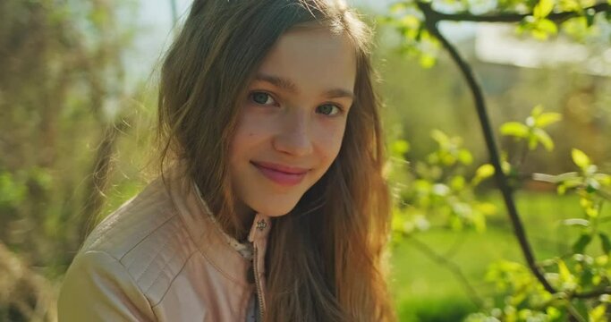 Preteen girl 10s has long blond hair, using mobile phone in the park and smiling at camera. Closeup portrait