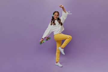 Fototapeta na wymiar Active teenager moves on purple background and holds longboard. Young girl in yellow pants and white hoodie listens to music in headphones.