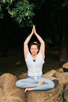 Yoga and Meditation. Happy young woman practicing meditation, sitting on a stone in yoga asana in the morning outdoors. Portrait of a smiling yogi woman exercising at sunrise. Vertical photo
