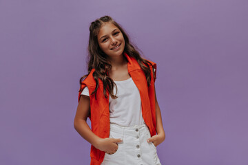 Beautiful curly girl smiles and looks into camera on purple background. Attractive tanned teenager in white skirt and orange sweater on shoulders poses.