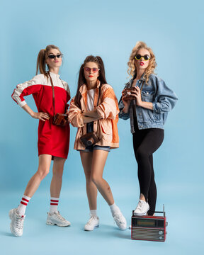 Pretty young women in retro 90s fashion style, outfits posing isolated over blue studio background. Concept of eras comparison, beauty, fashion and youth.