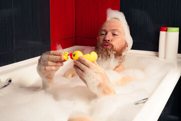 Cute funny bearded man playing with rubber ducks in bubble bath relaxing at home