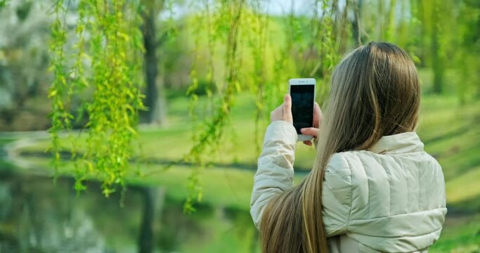 Preteen girl shooting photo with mobile phone when sitting on the grass near pond in the park. Girl has long blond hair looking at camera
