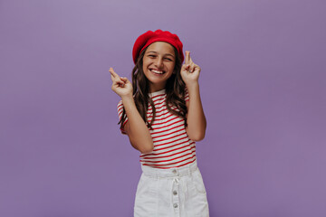 Obraz na płótnie Canvas Joyful curly girl in red beret points up at place for text on purple background. Excited teenager in striped t-shirt smiles on isolated.
