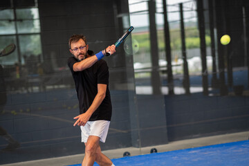 Man playing padel in a blue grass padel court indoor - Young sporty boy padel player hitting ball...