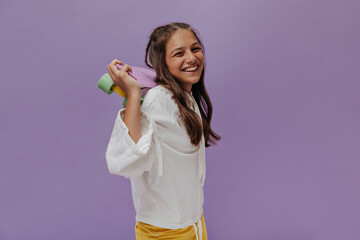 Charming young tanned girl in white hoodie smiles sincerely on purple background. Attractive teenager holds longboard.
