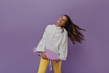 Good-humored tanned girl in white hoodie and yellow pants smiles sincerely, plays hair, holds longboard on purple background.