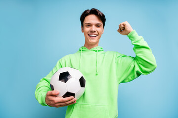 Photo of attractive lucky young gentleman wear green sweatshirt smiling holding ball rising fist isolated blue color background