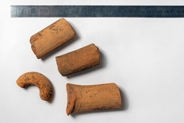 Some peices of clay aged-damaged ceramic artefacts found during the archaeological excavations settled on white sheet of paper, metallic ruler. Copy space, close up.